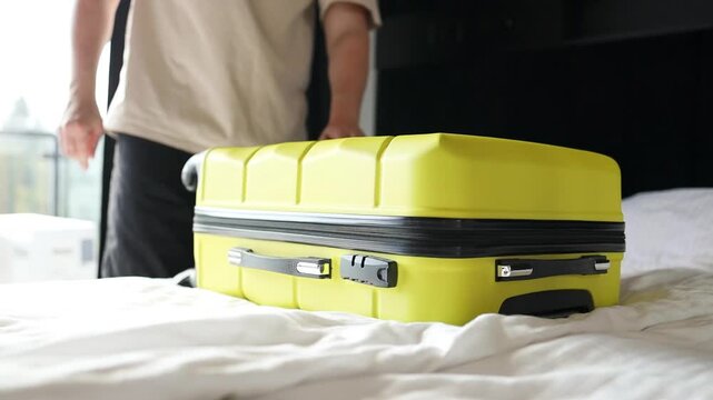 Close up of male hands carefully unzipping and opening a bright yellow hard-shell suitcase on a white bed. Unpacking clothes and baggage after vacation, arriving at a hotel room, or returning home
