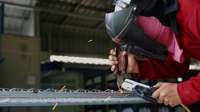 Welder wearing a protective face shield and gloves welds a steel rebar grid on the ground, with sparks and workshop tools visible in the background. Represents construction, metalwork, and industrial