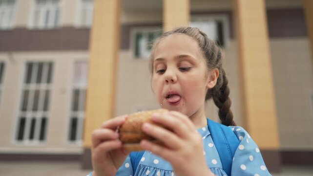Schoolgirl eats burger near school building. Child bites hamburger during lunch break. Kid enjoys fast food snack outdoors. Hungry pupil with backpack has quick meal. Student chews sesame bun.