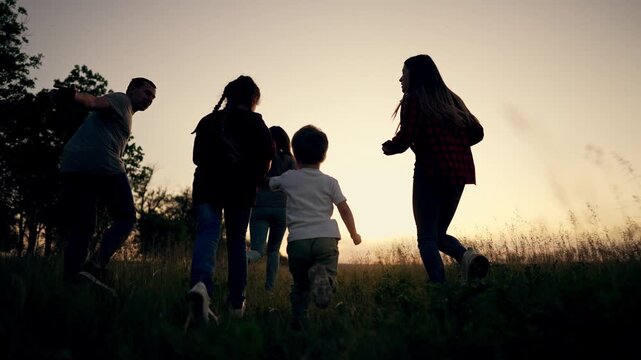 Happy family runs through sunset meadow with arms spread like airplanes. Parents and kids play freely in golden grass field. Silhouette of family bonding outdoors during summer evening at dusk.