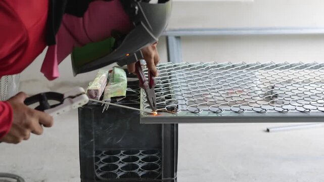 Welder wearing a protective face shield and gloves welds a steel rebar grid on the ground, with sparks and workshop tools visible in the background. Represents construction, metalwork, and industrial