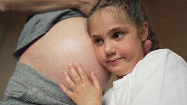 Child listens to mother pregnancy belly. Smiling sister hugs pregnancy belly. Sweet child touches mother bare skin. Future sister shares true family bonding love. Happy family awaits new baby.