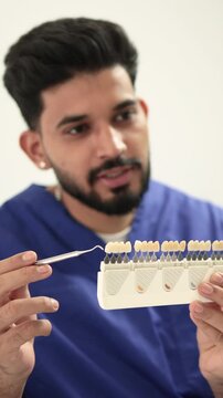 A male dentist in a blue uniform holds a tooth shade guide and a dental tool while consulting with a female patient