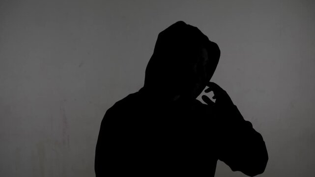 An anonymous silhouette of a man in a hood sits against a white, dirty wall. The man is giving an interview, actively gesturing with his hands and clutching his head in despair