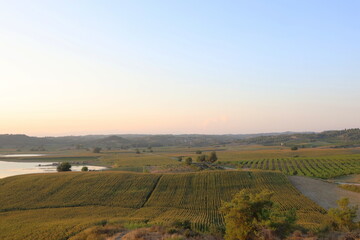 Fototapeta premium High-angle panoramic view of agricultural fields and hills next to a calm lake under a soft sunset sky.