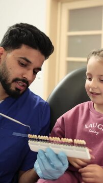 A male dentist in blue scrubs and gloves shows a young girl a tooth shade guide for dental procedures