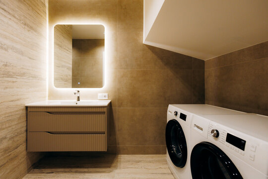 Modern laundry bathroom with illuminated mirror, floating vanity, front load washer and dryer beneath sloped ceiling, warm neutral finishes and compact storage, with bathroom, laundry, washer, dryer