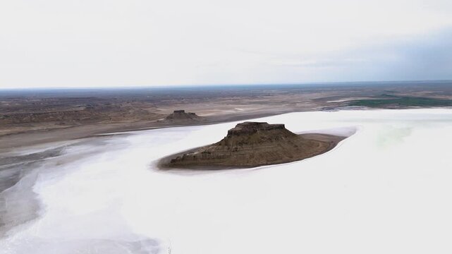 A smooth side aerial drone approach to a massive rock monolith standing alone in the misty, white-surfaced salt flat of Karynzharyk depression on a cloudy overcast day in Mangystau, Kazakhstan