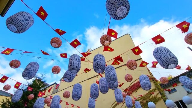 Paper lanterns and red decorative flags featuring yellow star and hammer and sickle symbols flutter in wind above courtyard outside. Low angle view against blue sky, festive cultural atmosphere.
