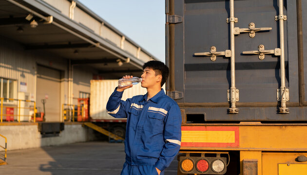 Young male logistics worker in blue uniform drinking water while leaning against a shipping container at sunset, hydration and break time for industrial labor and transport.