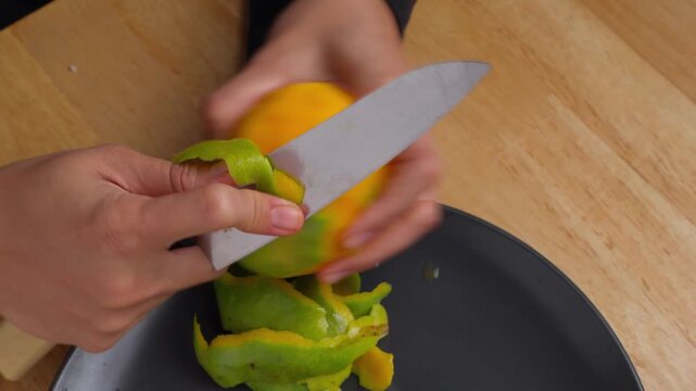 Hands peel mango with sharp knife over dark plate