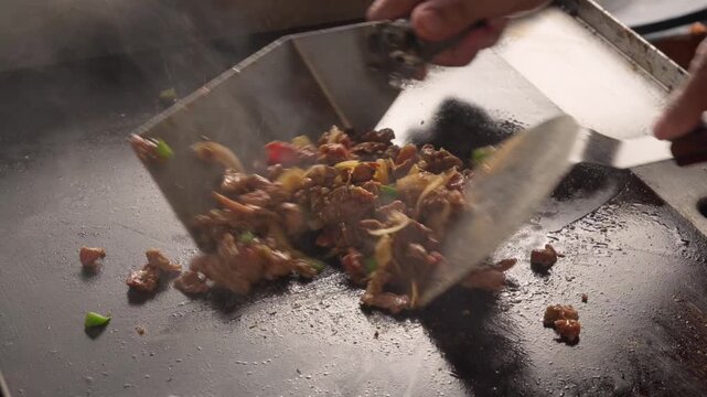Sizzling meat sizzles in a flat top hot griddle with onions and green chilies. Spatula flips the mixture, releasing steam and aroma