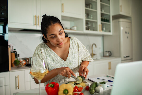 Young woman cutting cucumber while watching laptop in modern kitchen
