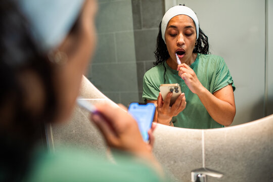 Young woman brushing teeth while using smartphone at bathroom mirror