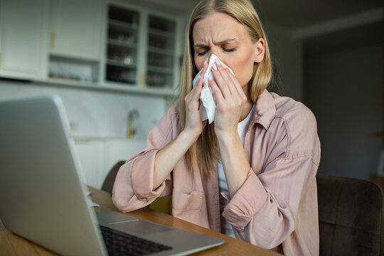 Sick woman blowing nose at laptop in home kitchen