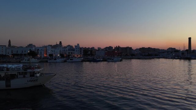Monopoli harbor reflecting a vibrant sunset sky over the water with boats moored and city lights illuminating the old town in apulia, italy, creating a serene scene