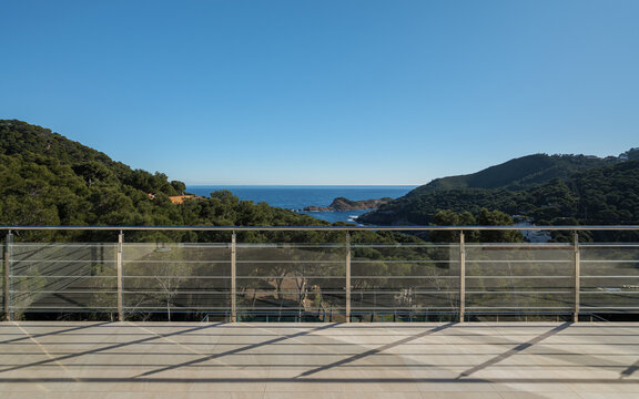Panoramic coastal view from terrace with glass railing overlooking rolling hills, blue ocean and distant horizon beneath clear sky in bright daylight, creating serene scenic setting