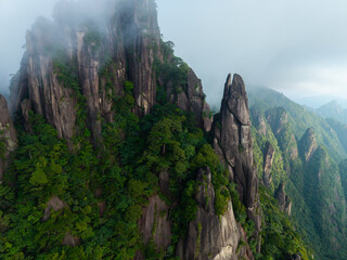 The majestic rocky peaks of Sanqingshan Mountain rise above the green forest landscape in the Jiangxi province of China surrounded by soft morning clouds and mist