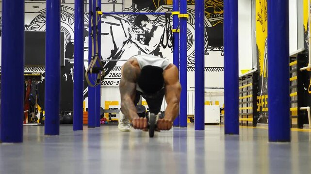 Strong black guy trying to do exercises with gymnastic wheel at modern gym. Muscular african sportsman practicing work with ab roller at fitness centre. Young afro american athlete training indoor