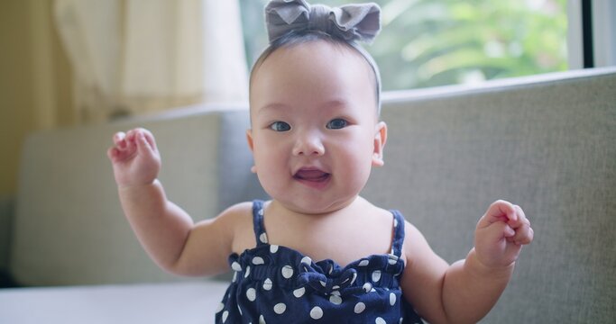 A happy Asian baby girl sits up on a sofa at home, looking at the camera with a curious and joyful expression, representing a candid moment of childhood innocence.