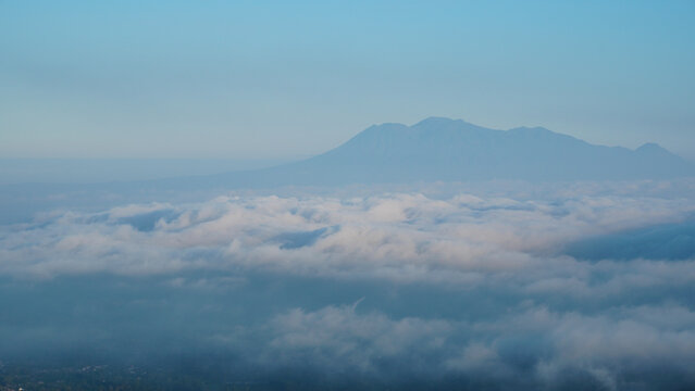 Telephoto view displays rolling white cloud bank hovering over unseen valley floor. Precise optical clarity captures distant mountain silhouette rendered in gentle blue dawn light.