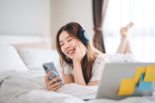 Mindfulness young adult asian woman lying in bed, wearing headphones listening relax music in cozy comfort bedroom