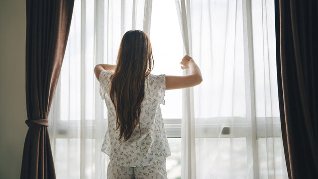 Rear view of young woman opening curtains in bedroom in the morning