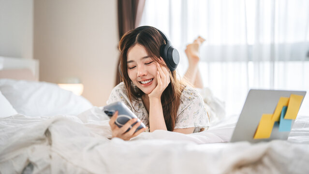 Mindfulness young adult asian woman lying in bed, wearing headphones listening relax music in cozy comfort bedroom