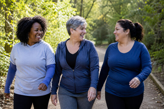 Three diverse midlife women walking and laughing together on a leafy park trail, showing friendship, social support, active aging, fitness, and menopause wellness.