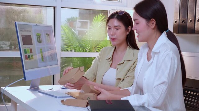 Two young asian woman designer working on packaging design with ai software with desktop computer, business team discussion of eco friendly cardboard box product development at modern home office.