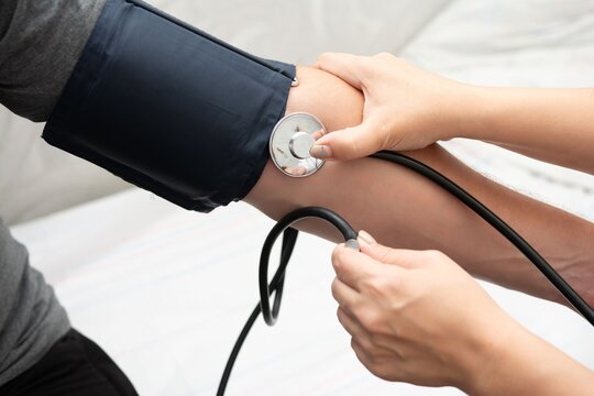 Healthcare provider using stethoscope and blood pressure cuff on a patient's arm to measure blood pressure in medical setting.