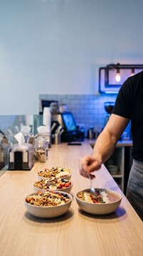 Chef in black shirt arranging granola smoothie bowls with fresh fruit toppings on wooden cafe counter, healthy breakfast trend and food service concept.