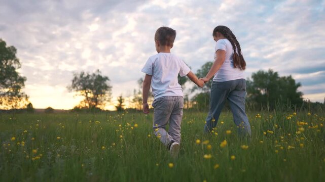 Mother and son walking through field. Girl is holding her hands and a boy is walking. Siblings playing in a field of grass at sunset. A mother and her son strolling across a meadow lifestyle.