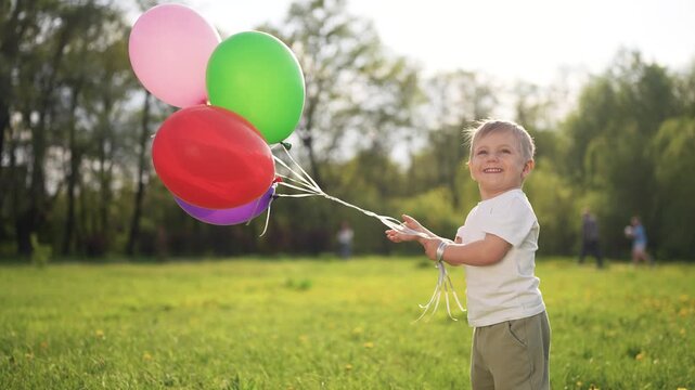 Young boy holding bunch of balloons. Colorful child holding son on run in the grass. A child is running with balloons in a lifestyle park. A little boy grasping a cluster of balloons.
