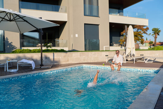 Man Relaxing by Modern Apartment Pool with Laptop While Someone Splashes and Swims