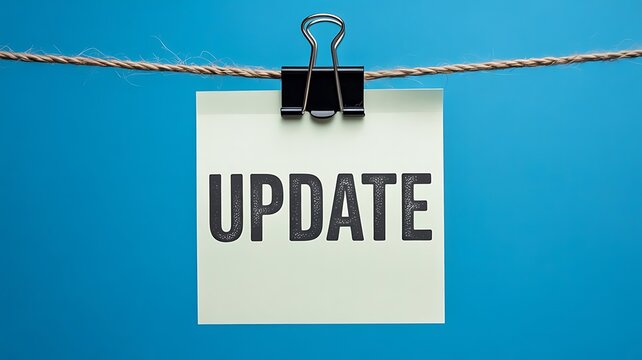 A white square sticky note with the word update printed in bold black letters hanging from a brown string with a black metal binder clip on blue