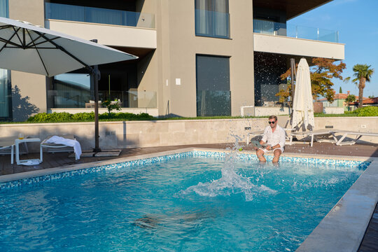 Man Sitting by Luxury Swimming Pool Splashing Water Outside Modern Villa on Sunny Vacation Morning