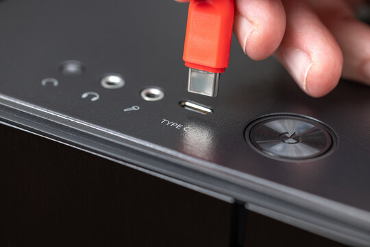 A hand inserts a red USB-C cable into the top panel of a desktop computer. The TYPE C label, metal mini jack audio ports, and round power button are visible. Connecting computer peripherals.