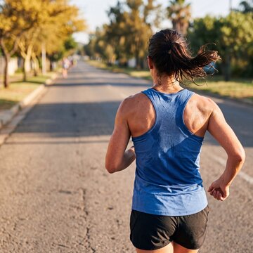 Back view of young woman in blue tank top jogging on sunny tree lined suburban street, morning run training and healthy active lifestyle concept.