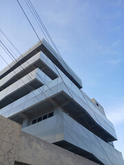 Fototapeta premium Low angle view of a sleek modern commercial building facade with metallic paneling and geometric architectural lines set against a clear blue sky during a bright and sunny afternoon day.