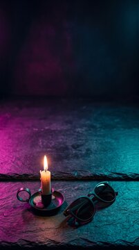 Burning candle in black metal holder and dark sunglasses on rough slate surface under neon pink and teal glow with moody vertical copy space.