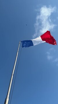 A French flag waving against a clear blue sky. The flag features blue, white, and red vertical stripes. Fluffy clouds are visible in the background.