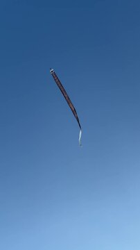 A long, thin object is soaring through a clear blue sky. The object appears to be a kite or a similar flying device, showcasing vibrant colors against the backdrop.