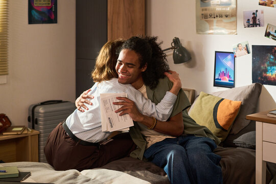 Young adult student hugging parent while holding accepted letter in bedroom, celebrating college admission and family support. Ideal for education success, future planning, medium shot