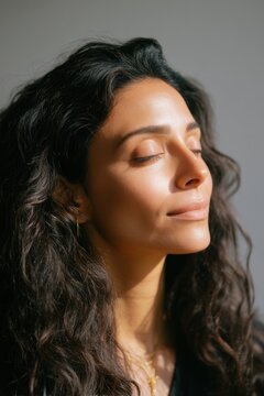 Beautiful woman with long dark wavy hair standing in sunlight. Portrait of female with glowing skin and closed eyes. Concept of wellness, self care, natural beauty and relaxation. Skincare.