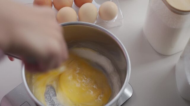Hand whisks eggs and sugar in metal saucepan on kitchen counter. Concept of cake batter preparation and homemade baking
