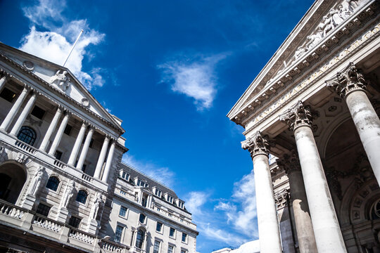 City of London financial district. Historic neoclassical pillars frame the Bank of England exterior under a clear blue sky.
