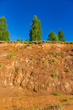Low angle view of a weathered sandy quarry slope with textured soil layers, sparse greenery, and green trees against a vibrant clear blue summer sky in daylight