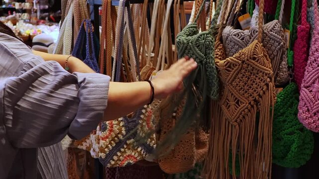 Woman choosing handmade macrame handbag in craft store. Female customer examining woven boho shoulder bag while shopping for artisan fashion accessories and handmade decor