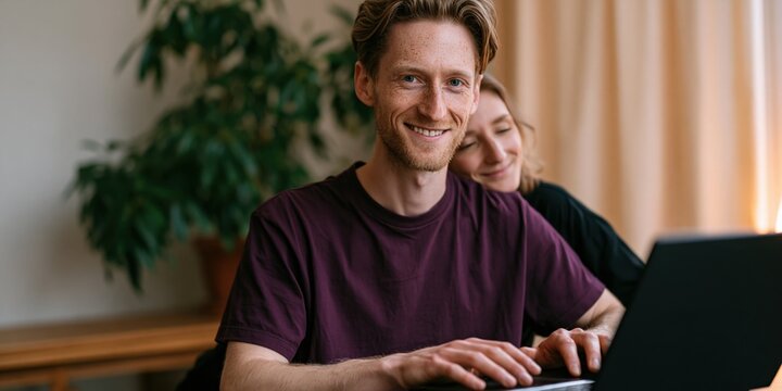 Young man typing on a laptop with partner leaning behind in a cozy home office for remote work background and productivity banner design